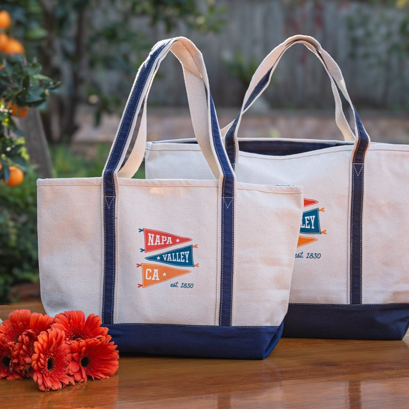 Two boat tote bags with Pennant Collection on a wooden surface with flowers and an orange tree in the background.