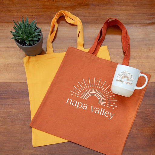 Orange tote bag with Sunshine 'Napa Valley' design, yellow tote bag, and white mug Terra Mug on a wooden surface.