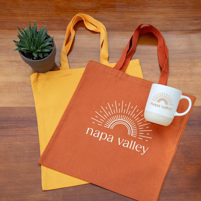 Orange tote bag with Sunshine 'Napa Valley' design, yellow tote bag, and white mug Terra Mug on a wooden surface.