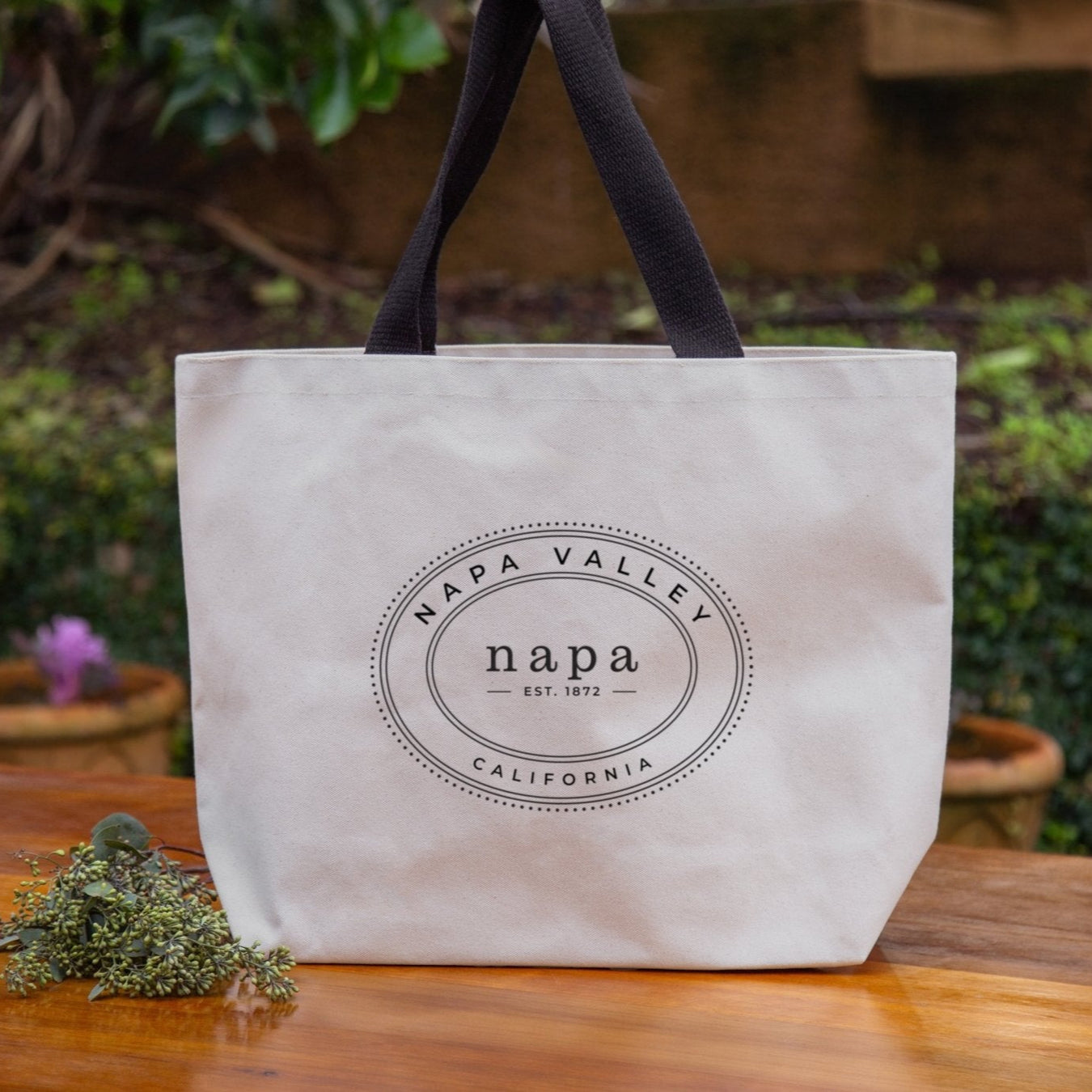 A beige heavyweight canvas tote bag with black cotton webbing straps, printed with Oval Collection 'Napa Valley' and 'California' on the front, placed on a wooden surface with foliage in the background.