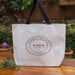 A beige heavyweight canvas tote bag with black cotton webbing straps, printed with Oval Collection 'Napa Valley' and 'California' on the front, placed on a wooden surface with foliage in the background.