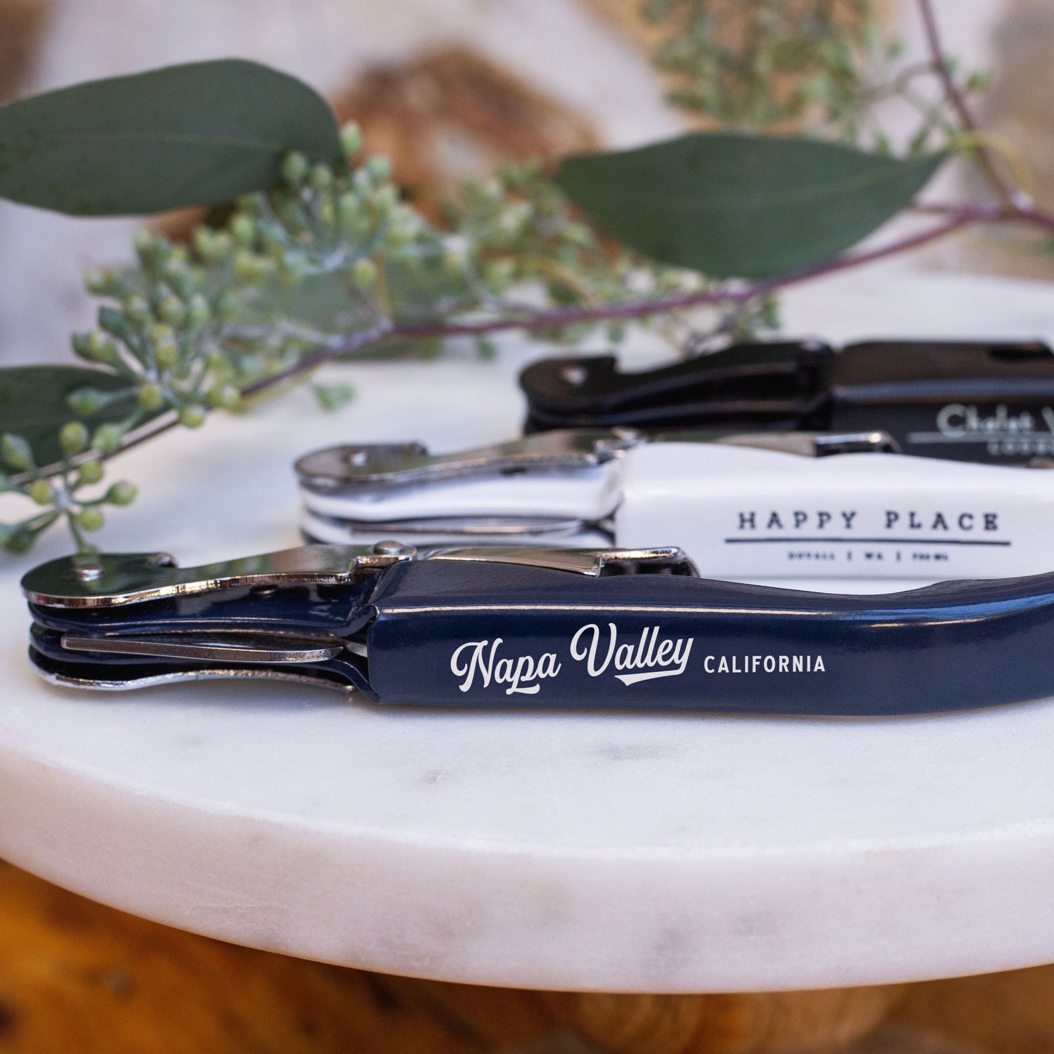 Set of wine bottle openers with 'Napa Valley' and 'Happy Place' branding on a marble surface.