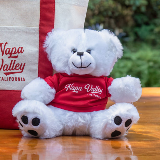 White teddy bear with a red 'Napa Valley' shirt sitting next to a matching tote bag on a wooden table outdoors.