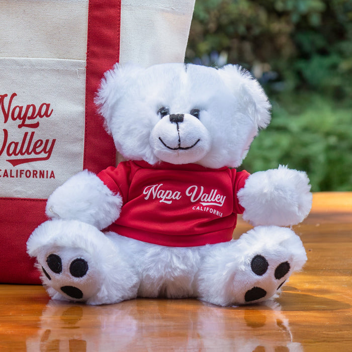 White teddy bear with a red 'Napa Valley' shirt sitting next to a matching tote bag on a wooden table outdoors.